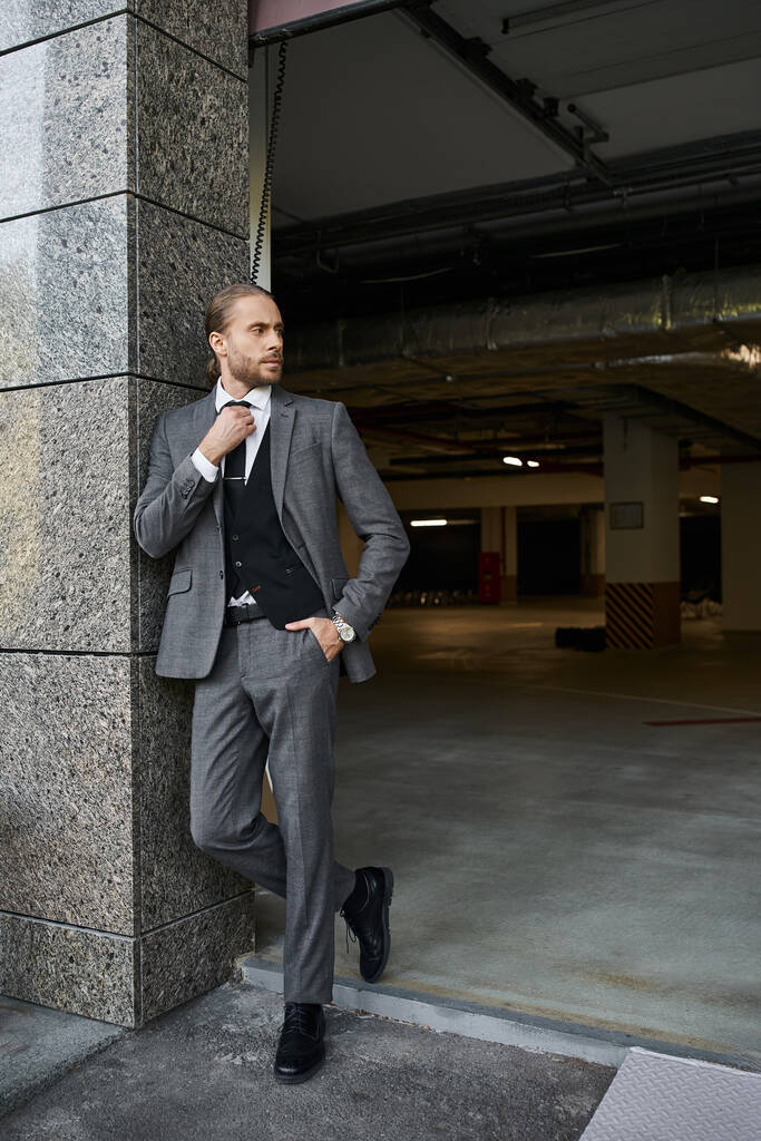 attrayant homme élégant avec barbe en costume intelligent gris posant à l'extérieur du parking, concept d'entreprise - Photo, image