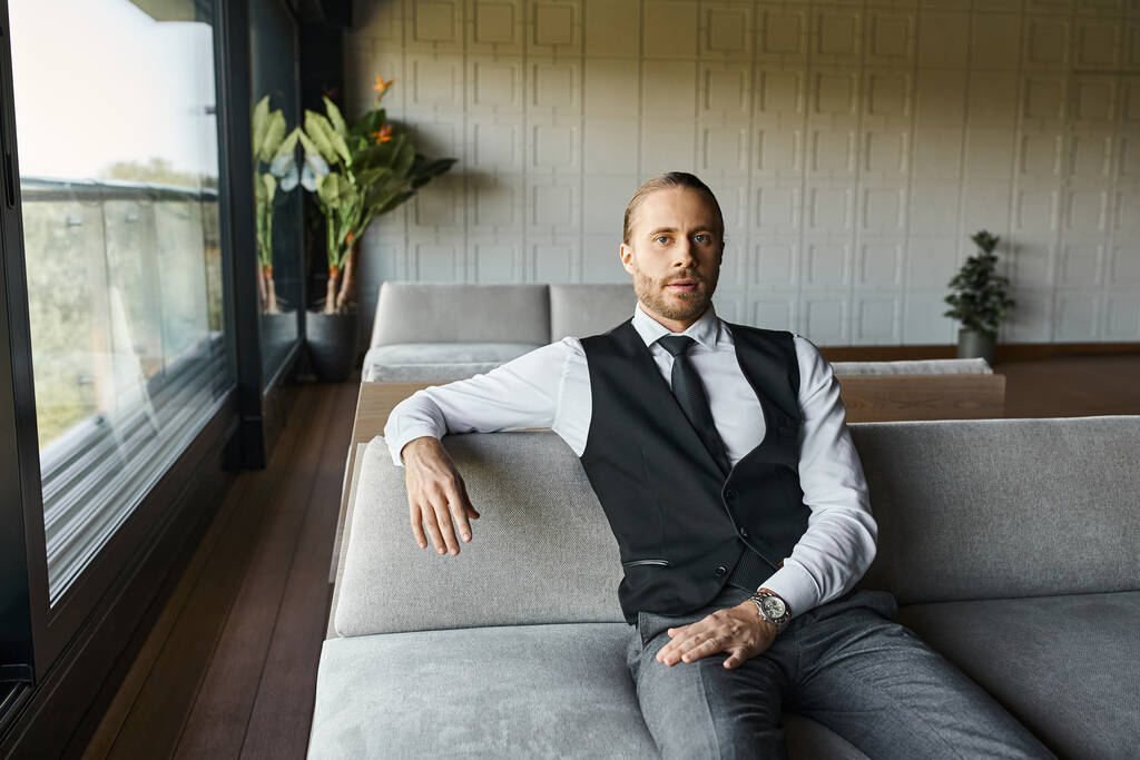 pohledný elegantní muž v černé elegantní vestě sedí na pohovce a dívá se do kamery, obchodní koncept - Fotografie, Obrázek