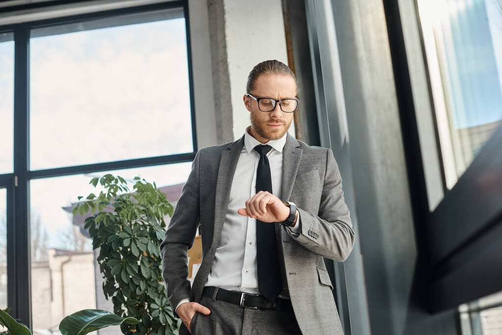 стильный бизнесмен в сером костюме и очках, стоящий в современном офисе и смотрящий на часы - Фото, изображение