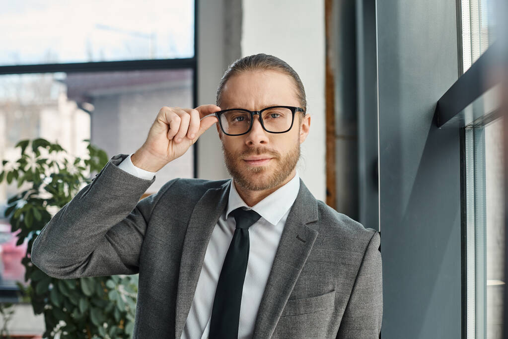 portrait of entrepreneur in formal wear adjusting eyeglasses and looking at camera in office - Photo, Image