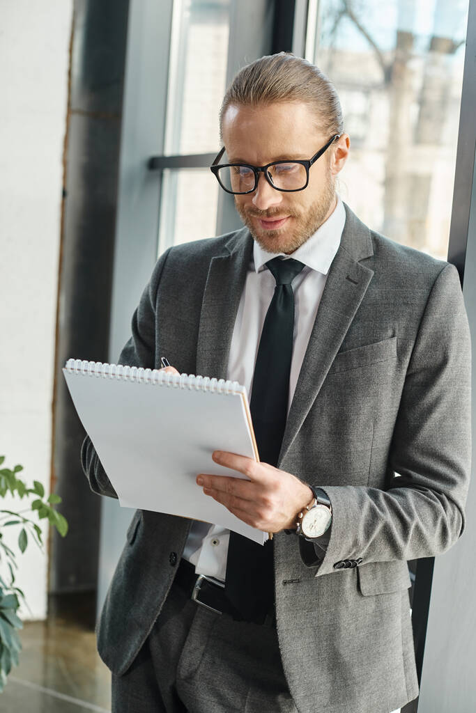 усміхнений бізнесмен у сірому костюмі та окулярах пише в блокноті, стоячи в офісі - Фото, зображення