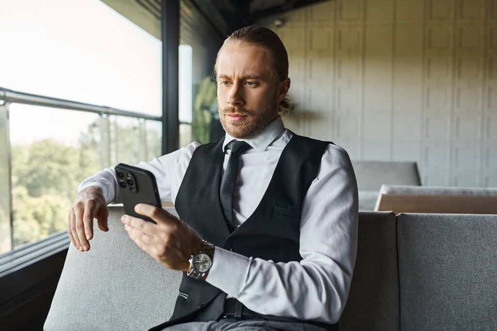 hombre de pelo rojo guapo en chaleco inteligente sentado en el sofá y mirando el teléfono móvil, concepto de negocio - Foto, Imagen