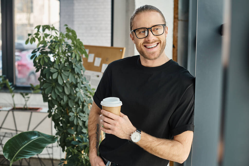 Siyah tişörtlü ve gözlüklü neşeli iş adamı elinde kahve tutarak ofisten dışarı bakıyor. - Fotoğraf, Görsel