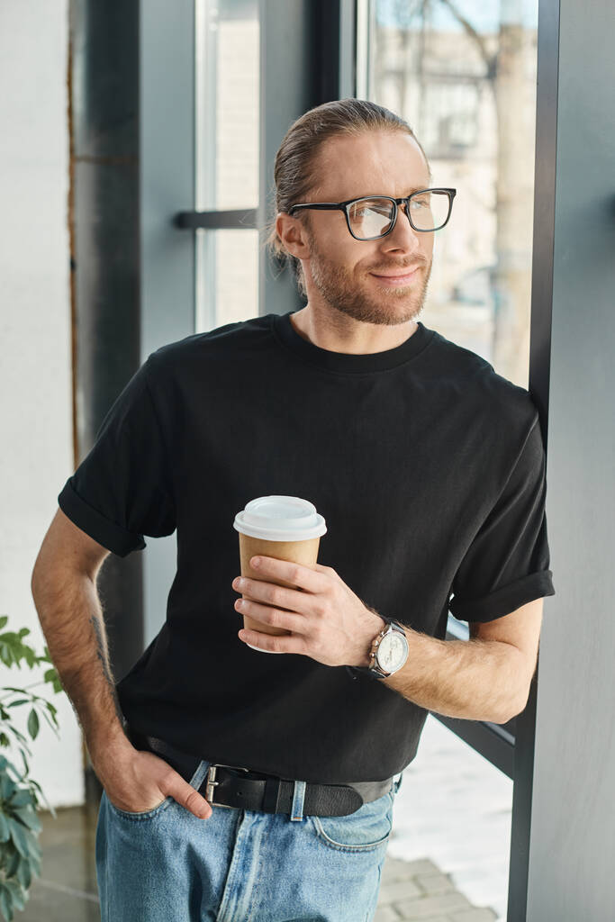 smiling manager in black t-shirt and eyeglasses holding takeaway drink and looking away in office - Photo, Image
