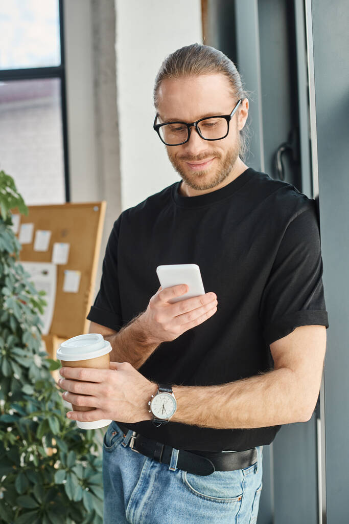 Gözlüklü ve günlük giysileri olan mutlu iş adamı akıllı telefona bakıyor ve elinde kahve tutuyor. - Fotoğraf, Görsel