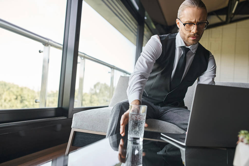 красивый элегантный бизнесмен в элегантной одежде с галстуком, работающий на ноутбуке, поднимая стакан с водой - Фото, изображение