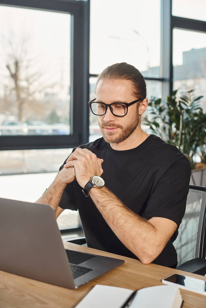 вдумчивый бизнесмен в черной футболке и очках смотрит на ноутбук на рабочем месте в офисе - Фото, изображение
