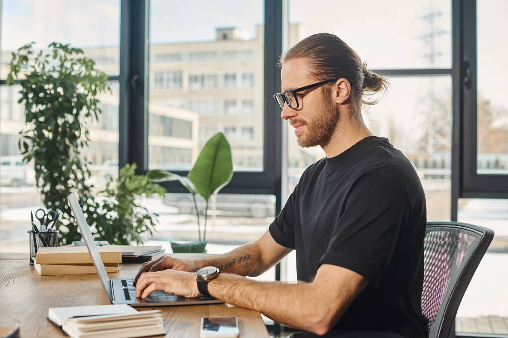 uomo d'affari sorridente in t-shirt nera e occhiali da vista che digita sul computer portatile sul posto di lavoro in ufficio moderno - Foto, immagini