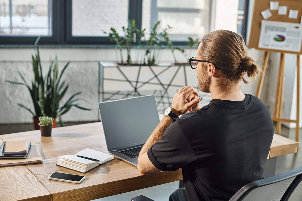 Rückansicht des Managers in schwarzem T-Shirt neben Laptop mit leerem Bildschirm am Schreibtisch im modernen Büro - Foto, Bild