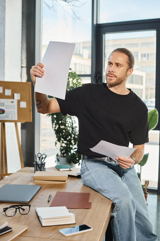 serious businessman in casual clothes sitting on work desk near and paper in modern office - Photo, Image