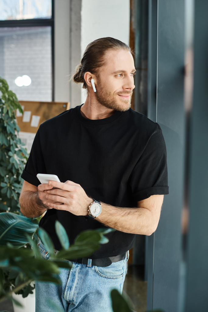 χαρούμενος επιχειρηματίας με μαύρο t-shirt κρατώντας smartphone και ακούγοντας μουσική στα ακουστικά στο γραφείο - Φωτογραφία, εικόνα