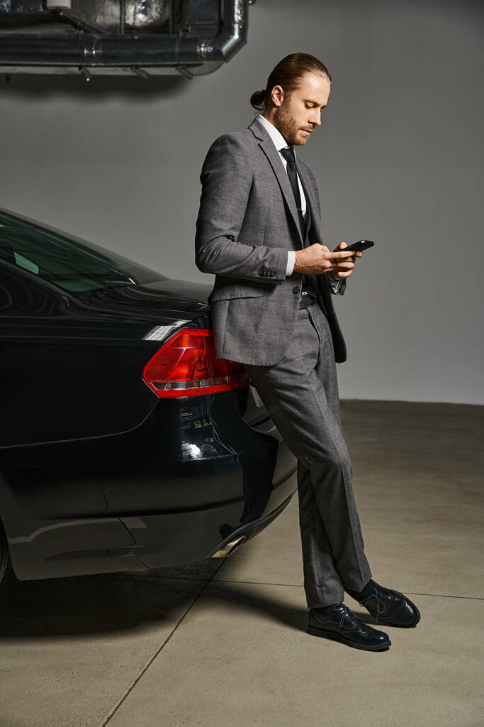 pohledný elegantní podnikatel v obleku s rudými vlasy opírající se o jeho auto a při pohledu na telefon - Fotografie, Obrázek