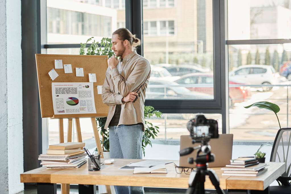 στοχαστικός επιχειρηματίας κοιτάζοντας flip charts με infographics κοντά ψηφιακή φωτογραφική μηχανή στο γραφείο - Φωτογραφία, εικόνα