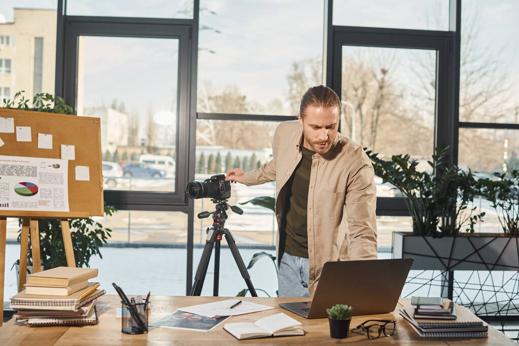 オフィスのワークデスク,ビデオブロガーの近くのラップトップとデジタルカメラを調整するカジュアルな服装のマネージャー - 写真・画像