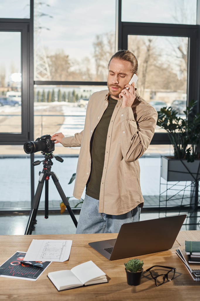 現代オフィスのデジタルカメラやラップトップの近くのスマートフォンで話すビジネスマン - 写真・画像