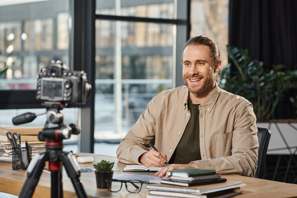 homme d'affaires heureux souriant à l'appareil photo numérique et l'écriture dans un cahier sur le lieu de travail dans le bureau moderne - Photo, image