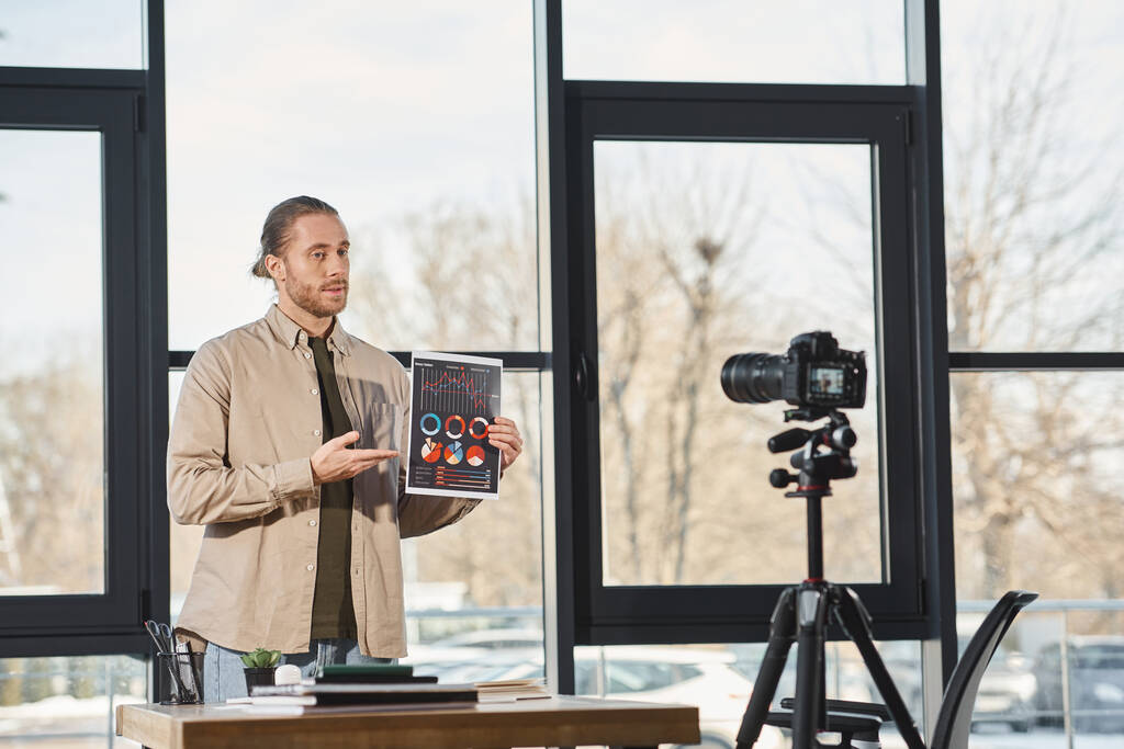 επιχειρηματίας δείχνει έγγραφο με infographics κατά τη διάρκεια βίντεο στο blog ψηφιακή φωτογραφική μηχανή στο γραφείο - Φωτογραφία, εικόνα
