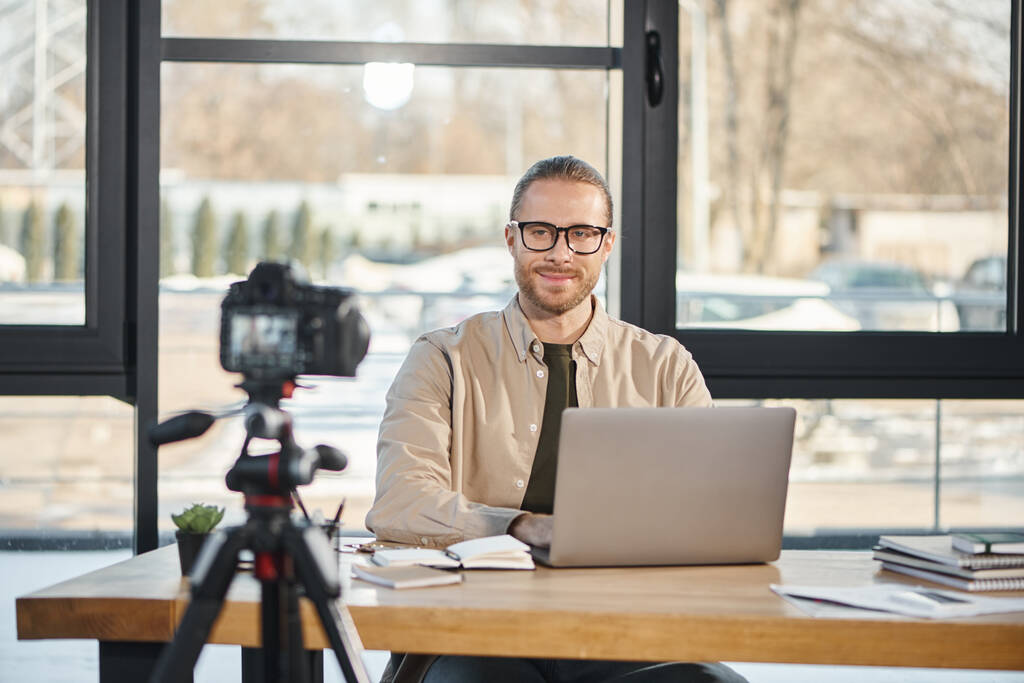 χαρούμενος διευθυντής σε γυαλιά ηλίου κάθεται κοντά στο laptop και χαμογελώντας στην ψηφιακή φωτογραφική μηχανή κατά τη διάρκεια βίντεο blog - Φωτογραφία, εικόνα