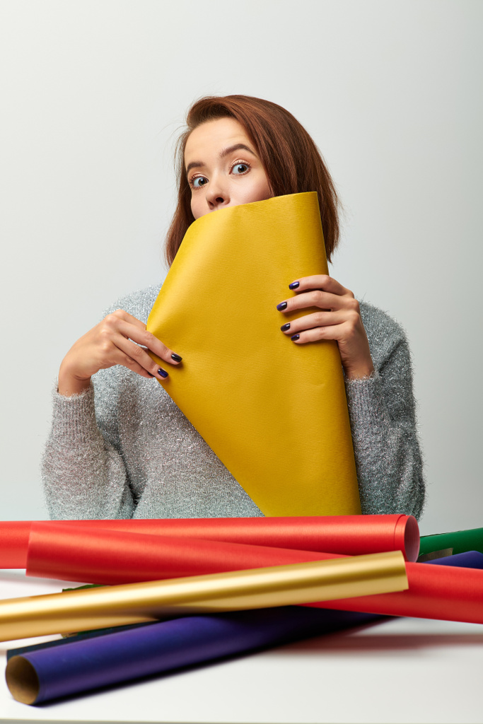 Weihnachtszeit, junge Frau im Pullover mit gelbem Geschenkpapier vor grauem Hintergrund - Foto, Bild