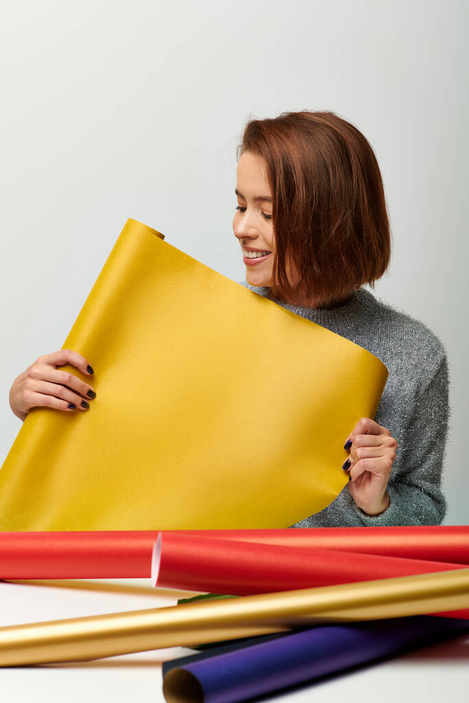 mosolygós fiatal nő pulóverben kezében sárga ajándék papír szürke háttér, Boldog Karácsonyt koncepció - Fotó, kép