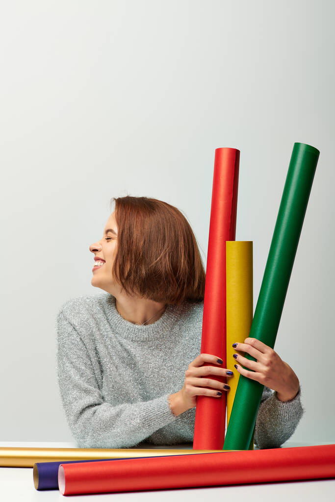 радісна жінка в затишному светрі тримає різні кольори подарункового паперу на сірому фоні, з Різдвом Христовим - Фото, зображення