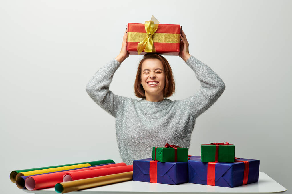 wesoła kobieta w zimowym swetrze trzyma prezent świąteczny nad głową przy papierze upominkowym na szaro - Zdjęcie, obraz
