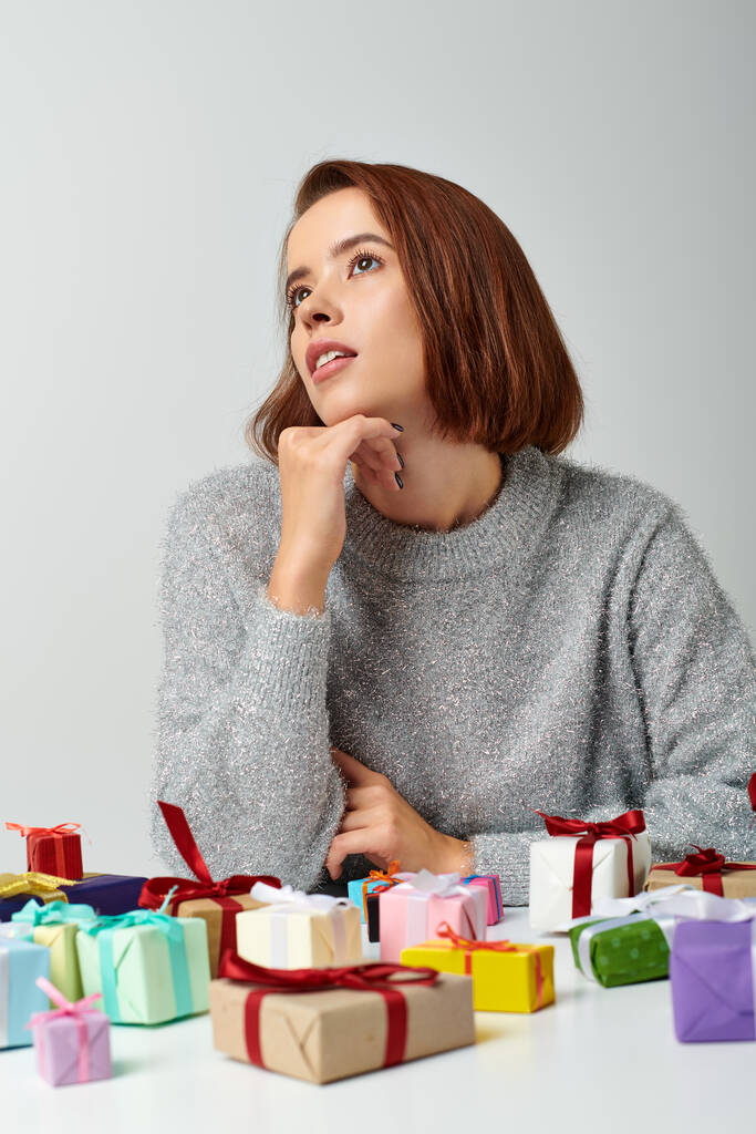 pretty woman in winter sweater fantasizing among bunch of Christmas gifts on table, grey backdrop - Photo, Image