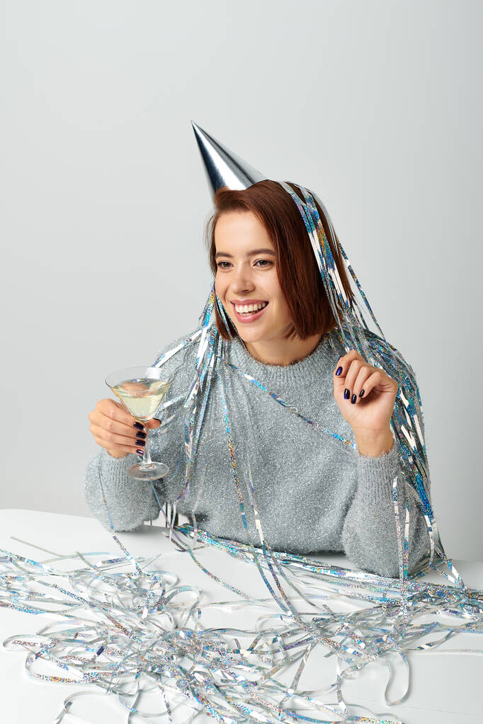 Frau in Partymütze mit silbernem Lametta auf dem Kopf hält Glas Champagner auf grau, Frohes Neues Jahr - Foto, Bild