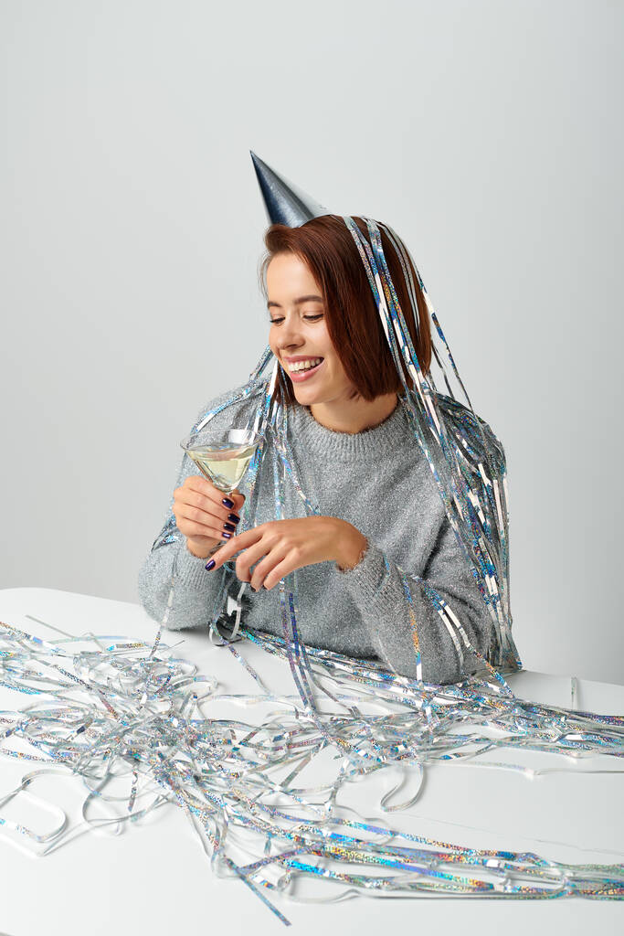 donna allegra in berretto partito con fili di lame sulla testa che tiene un bicchiere di champagne su grigio, felice anno nuovo - Foto, immagini