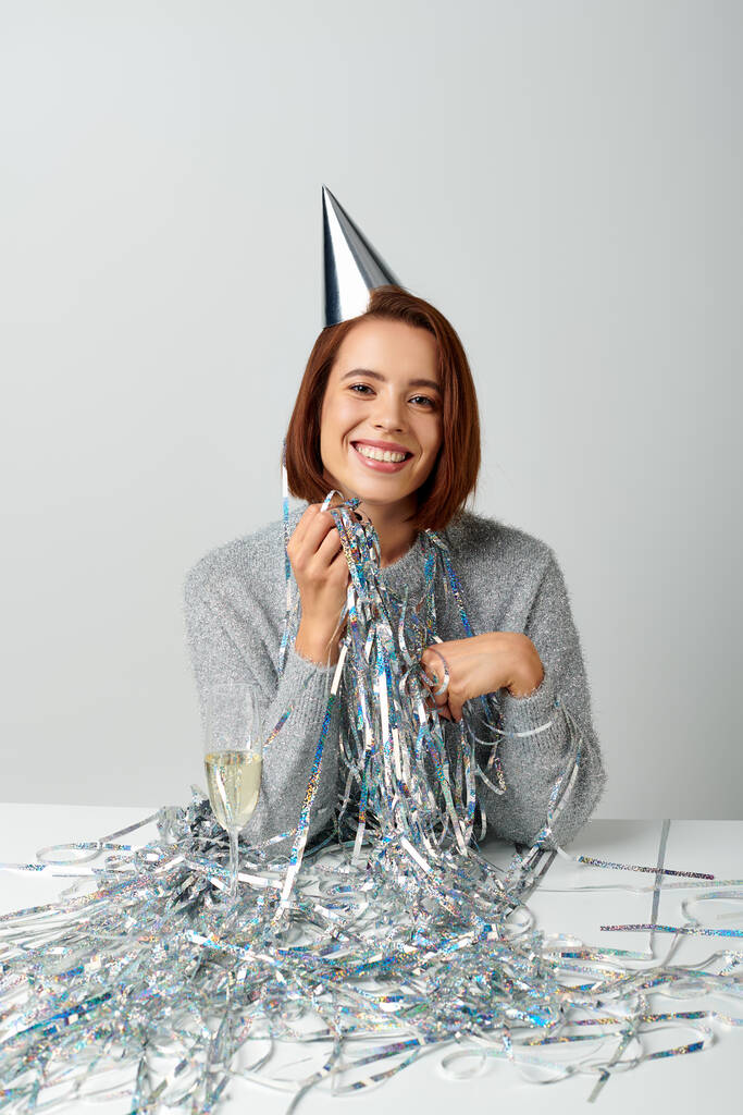 mujer contenta en gorra de fiesta y oropel sonriendo cerca de copa de champán mientras se celebra el Año Nuevo - Foto, imagen