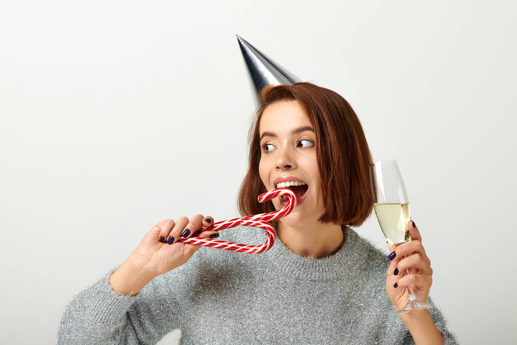 happy woman in party cap holding champagne glass and biting candy cane on grey, Merry Christmas - Photo, Image