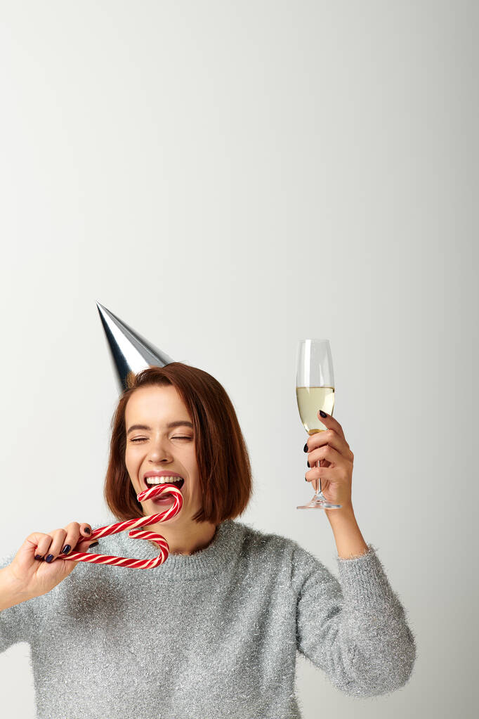 Parti şapkalı genç kadın elinde şampanya bardağı ve gri Noel 'de şeker kamışı ısırıyor. - Fotoğraf, Görsel