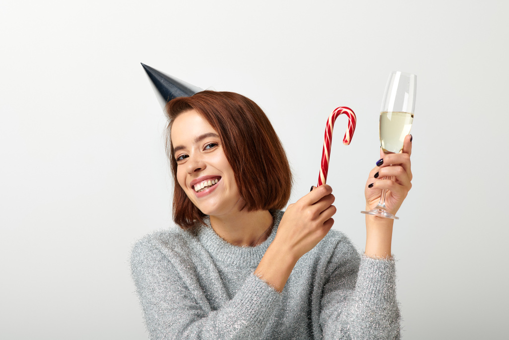 パーティーキャップの肯定的な女性は,グレー,メリークリスマスにシャンパンガラスとキャンディーケインを保持 - 写真・画像