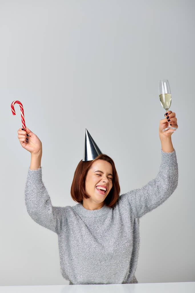opgewonden vrouw in party cap houden snoep riet en champagne glas op grijze achtergrond, Gelukkig Nieuwjaar - Foto, afbeelding