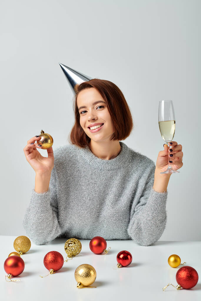 Frau mit Partymütze hält Champagnerglas und Christbaumkugel auf grau, Frohe Weihnachten Konzept - Foto, Bild