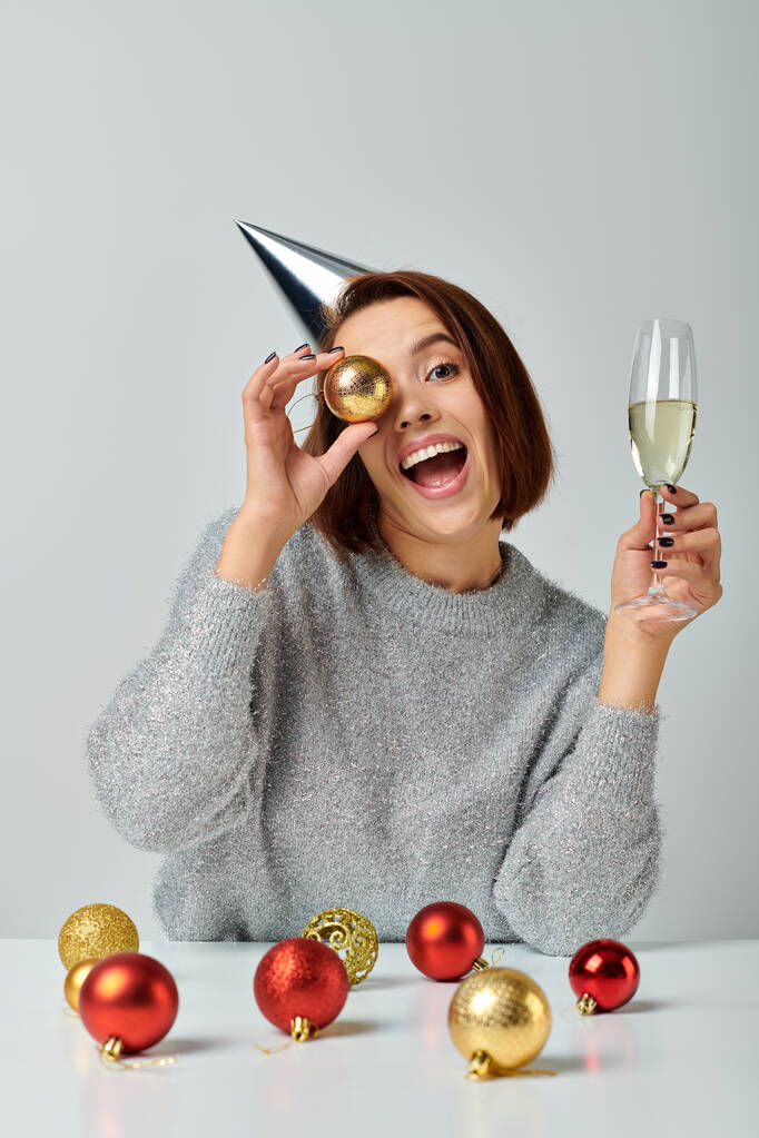 взволнованная женщина в кепке для вечеринок с бокалом шампанского и безделушкой на сером, с рождественской концепцией - Фото, изображение