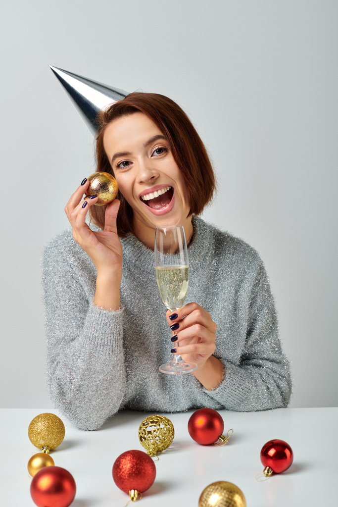 donna eccitata in berretto partito che tiene bicchiere di champagne e bauble di Natale sul grigio, Felice anno nuovo - Foto, immagini