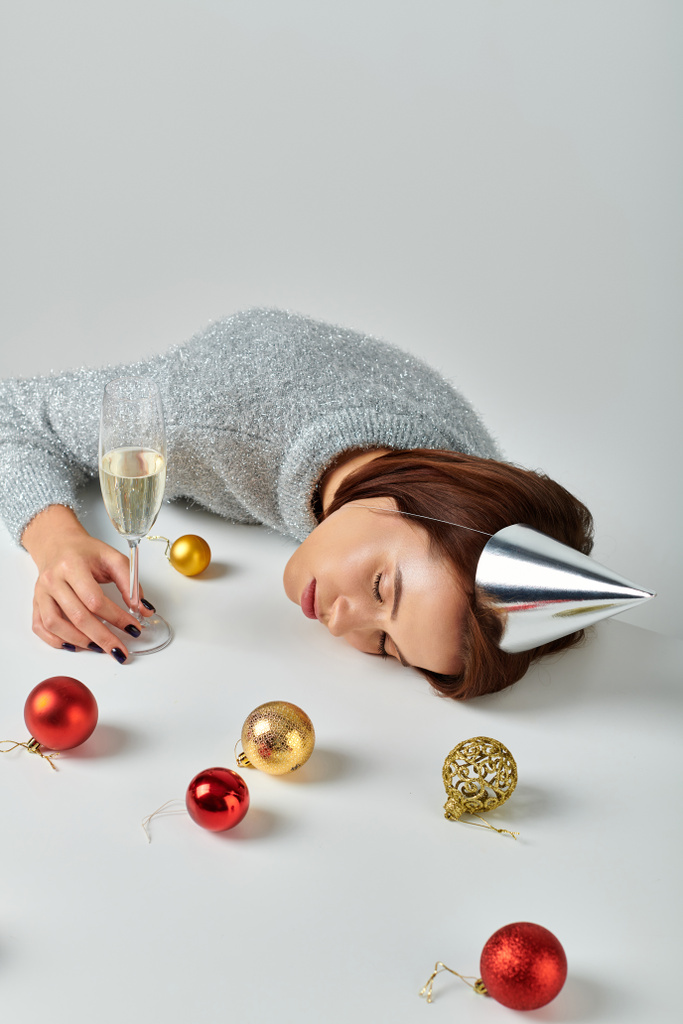 mujer con gorra de fiesta durmiendo en la mesa cerca de la copa de champán y adornos de Navidad sobre fondo gris - Foto, imagen