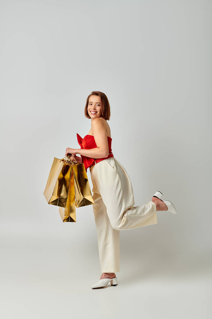 Kerstinkopen, vrolijke jonge vrouw in trendy kleding met boodschappentassen op grijze achtergrond - Foto, afbeelding