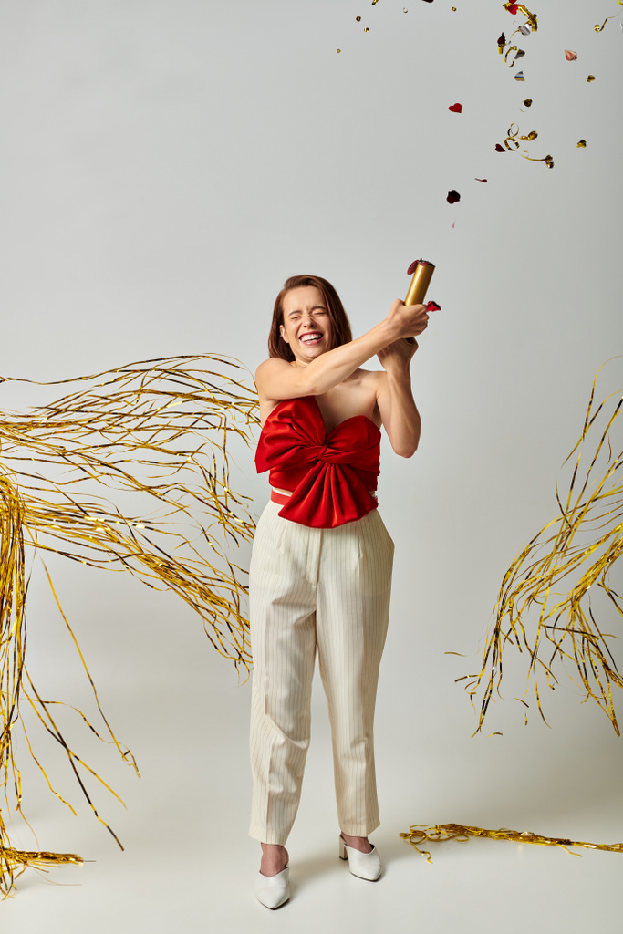 Feliz Ano Novo, alegre jovem mulher em traje na moda batendo palmas confete perto de ouropel no fundo cinza - Foto, Imagem