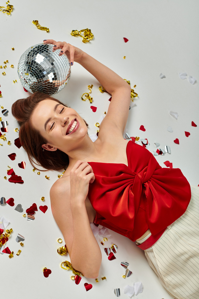 Festa de Ano Novo, jovem mulher alegre com bola de discoteca deitada no chão perto de confete em pano de fundo cinza - Foto, Imagem