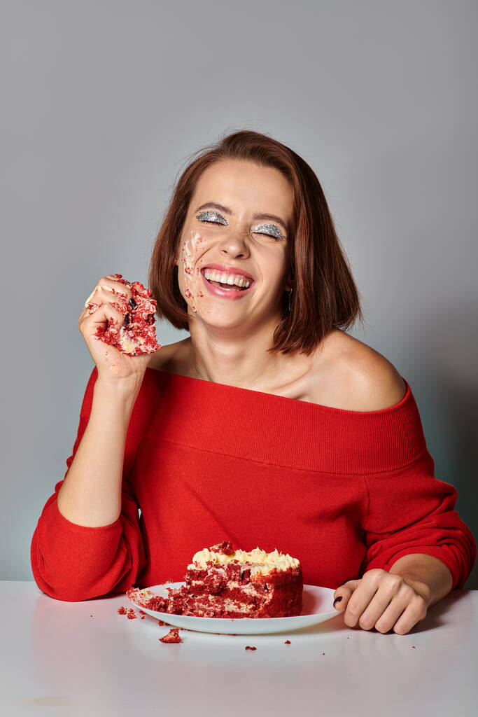 pozitif doğum günü kızı, kapalı gözlü, gri arka planda lezzetli bir parça kırmızı kadife kek yiyor. - Fotoğraf, Görsel