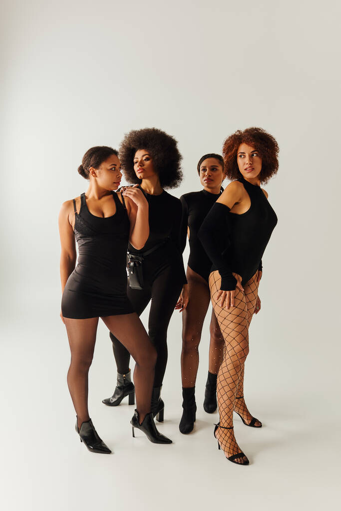 cuatro mujeres afroamericanas atractivas en traje atractivo negro posando juntas, concepto de moda - Foto, Imagen