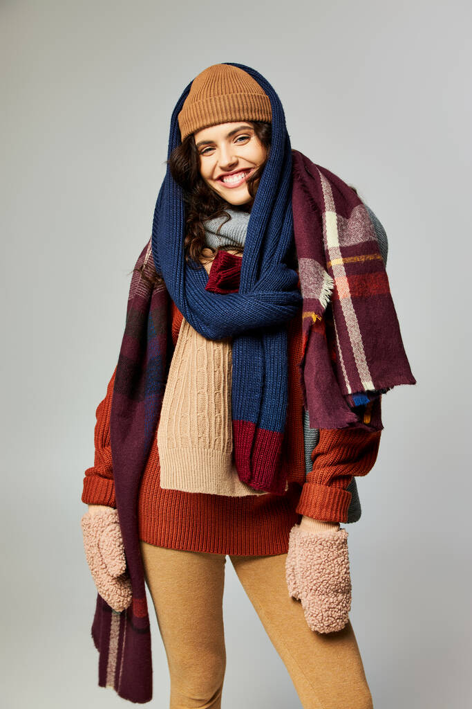 moda de inverno, modelo alegre em roupas em camadas, chapéu quente e lenços posando no fundo cinza - Foto, Imagem