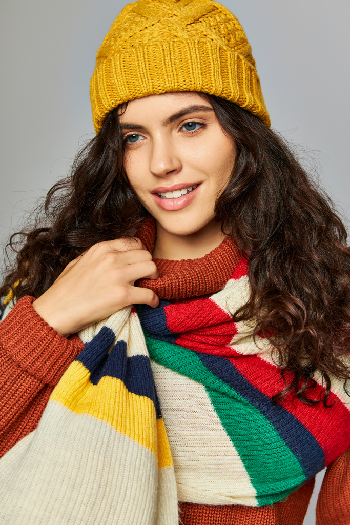 wesoła kobieta w zimowym kapeluszu bobble i sweter z rozebranym szalikiem pozowanie na szarym tle - Zdjęcie, obraz