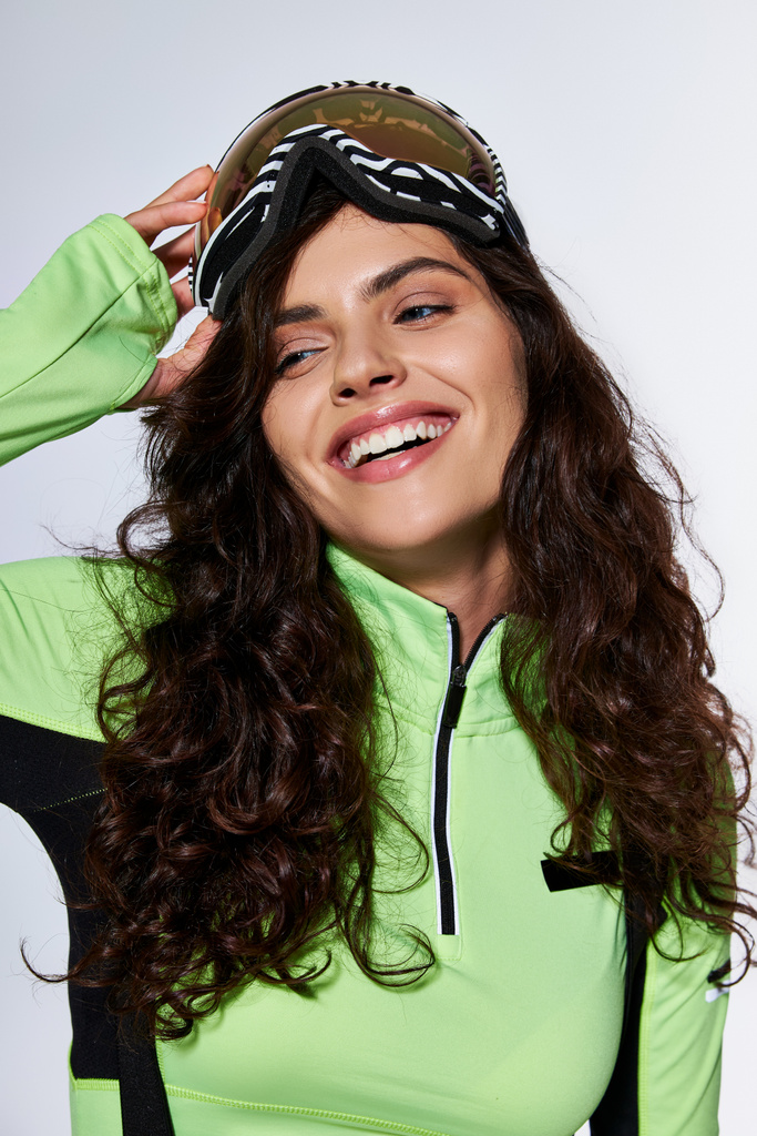 χαρούμενη νεαρή γυναίκα με σγουρά μαλλιά ποζάροντας σε κομψά ρούχα του σκι και γυαλιά σε γκρι φόντο - Φωτογραφία, εικόνα