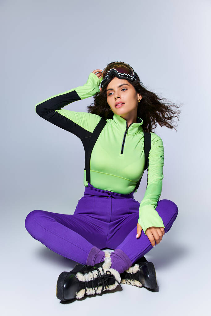 urheilullinen nuori nainen, jolla on kiharat hiukset, poseeraa trendikkäässä aktiivisessa kulumisessa istuessaan harmaalla taustalla - Valokuva, kuva