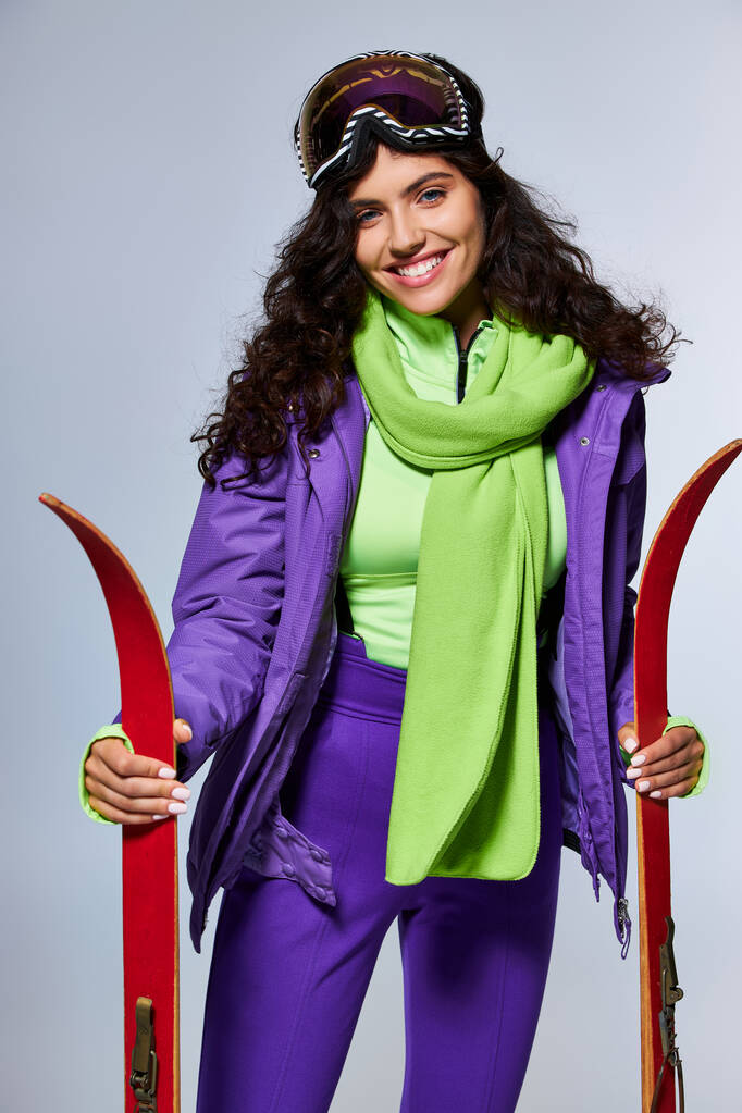 zimowy wypoczynek, pozytywna kobieta z kręconymi włosami pozująca w aktywnym ubraniu z kurtką i nartami - Zdjęcie, obraz