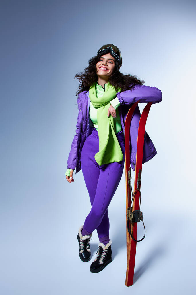 actividad invernal, mujer satisfecha con el pelo rizado posando en desgaste activo con chaqueta hinchable y esquís - Foto, Imagen