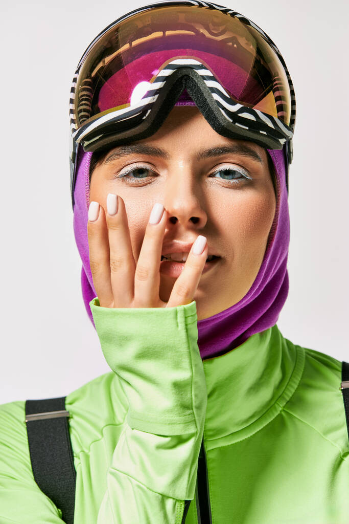 mujer joven en ropa de esquí de invierno con pasamontañas en la cabeza posando con las manos cerca de la cara en gris - Foto, Imagen